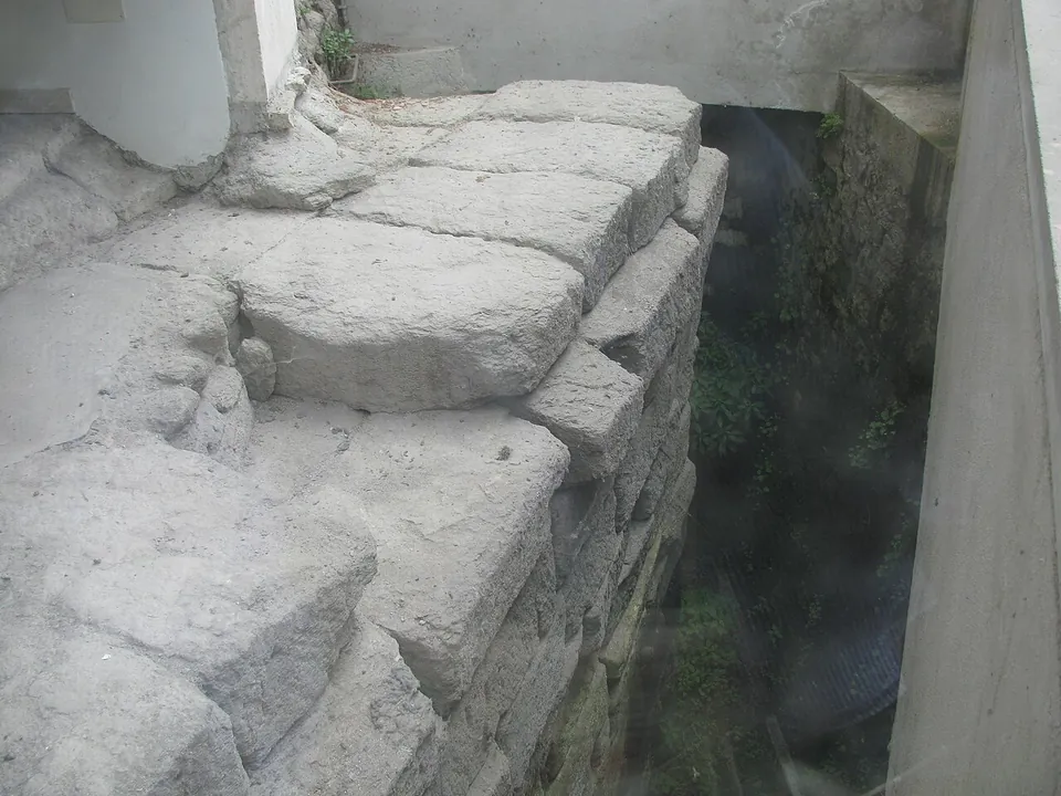 Close view of weathered grey stone blocks stacked at the edge of a deep shaft, with later masonry and plants in the shadow below.