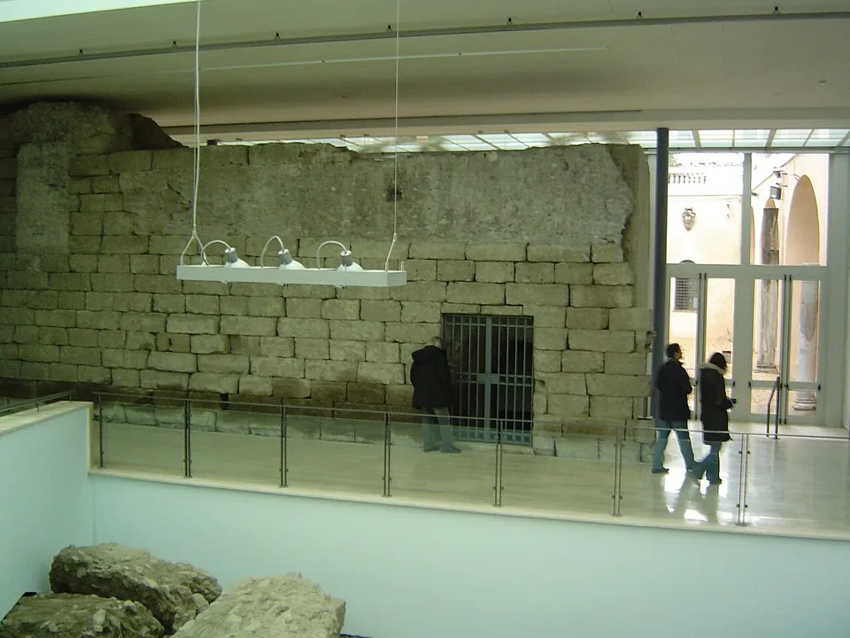 A long wall of squared grey stone blocks, partly broken along its top, running the length of a modern museum hall with a glass balustrade in front of it, a barred door set into its base and two visitors walking past on the right.