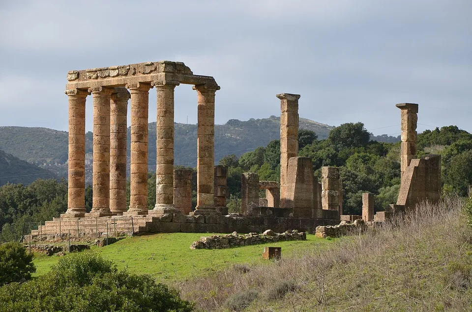 Tall columns of honey-coloured stone carrying a short stretch of entablature at the front of the temple, with stumps of pillars and walls standing on the platform behind them, a grassy slope in front and wooded mountains beyond.