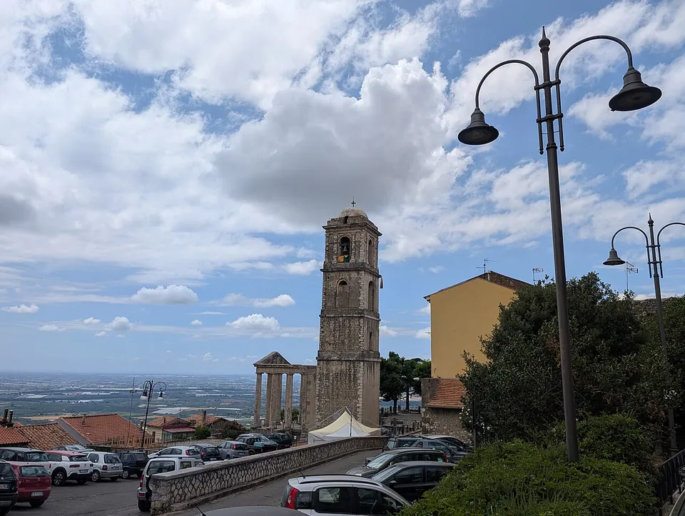 A square at the top of the town with parked cars, a low wall and a tall stone bell tower; beside the tower stands a small temple front of pale columns, with the flat plain stretching to the horizon behind.
