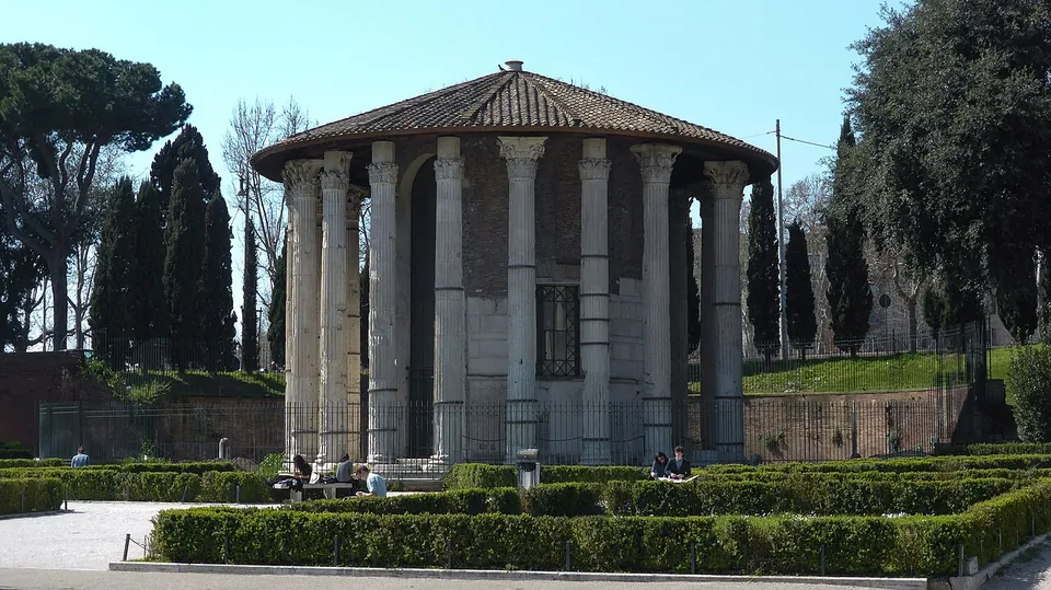 A round temple ringed by tall fluted columns with Corinthian capitals under a low conical tiled roof, standing behind a railing in a garden of clipped hedges, with people sitting on a bench in front.