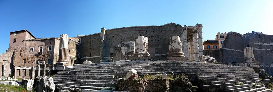 A panorama of a broad flight of stone steps rising to a ruined temple platform with broken column bases on it, three tall columns at the right, and a very high wall of grey stone blocks closing the view behind.