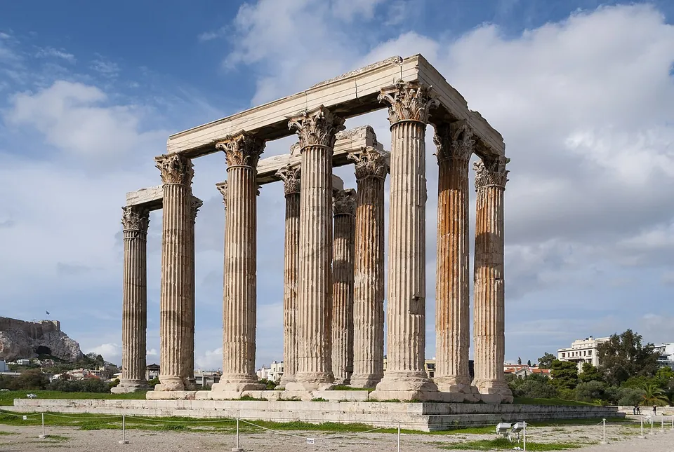 A group of colossal fluted marble columns with Corinthian capitals, still joined by stretches of their stone beams, standing on a low platform under a cloudy sky, with the Acropolis rock in the distance on the left.