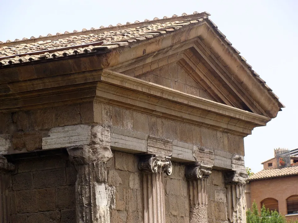 The upper part of a small stone temple, its tiled roof and plain triangular pediment above a cornice, with Ionic capitals on the free-standing column at the corner and on half-columns set into the wall beside it.
