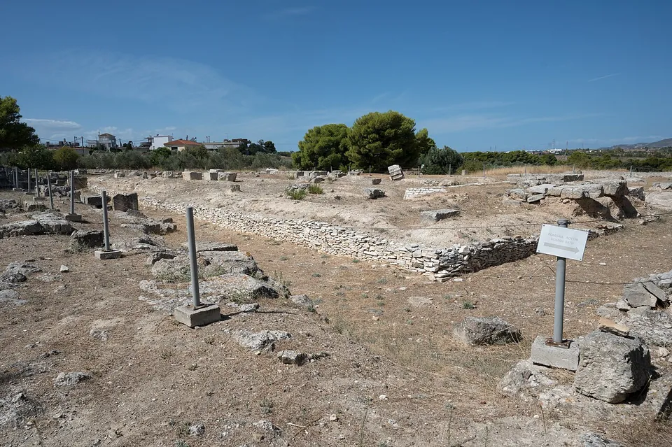A broad, flat excavated area of low foundations and scattered stone blocks on dry ground, with an information sign and a few trees and houses beyond.