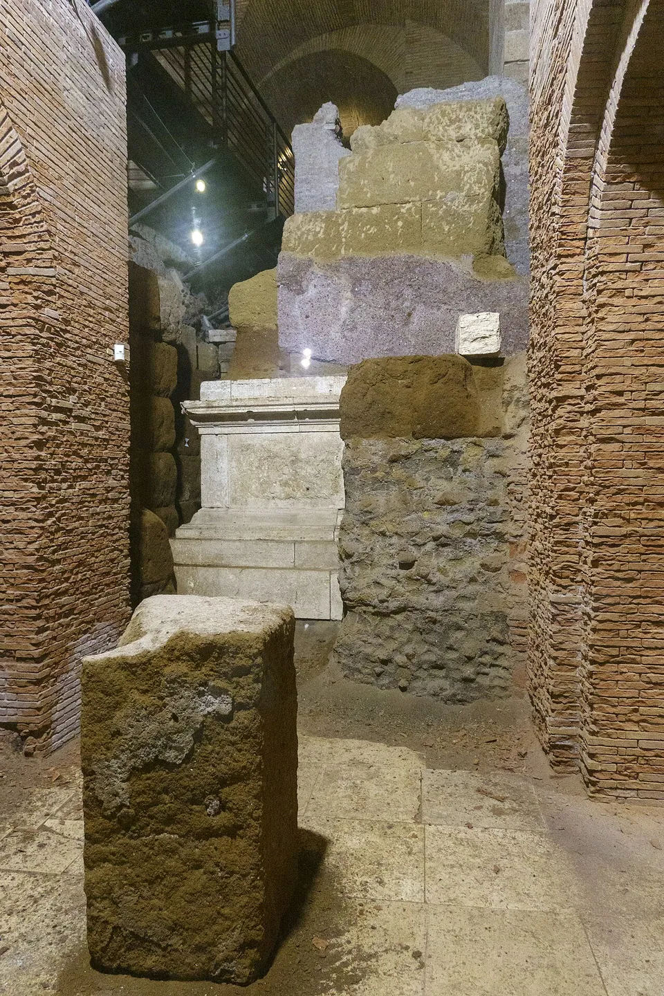 A narrow passage between brick walls under a vaulted ceiling, with blocks of travertine and tufa stacked in a rough stepped column at its centre.
