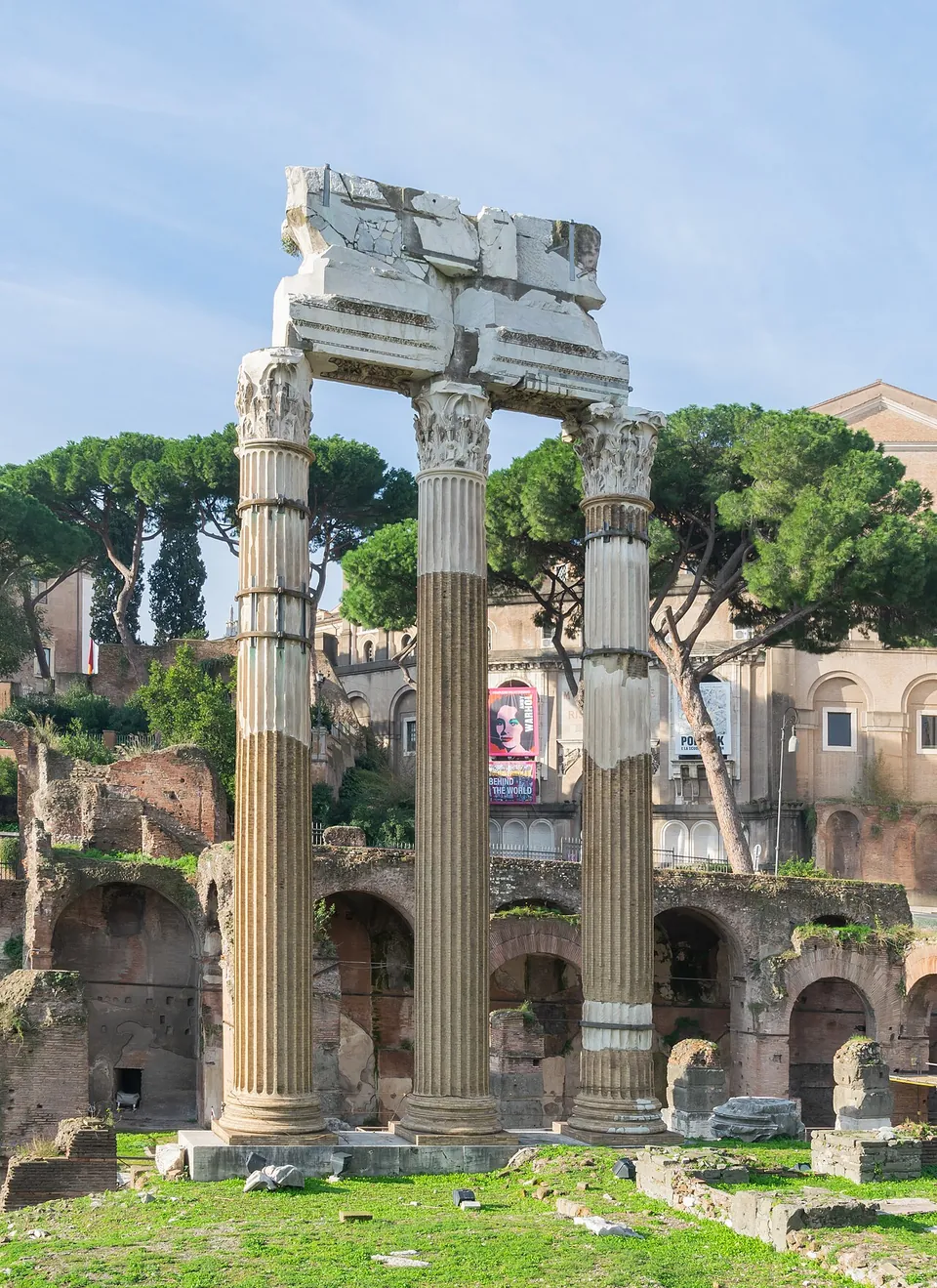 Three fluted columns with Corinthian capitals carrying a broken block of entablature, standing on a lawn in front of ruined brick arcades and umbrella pines.