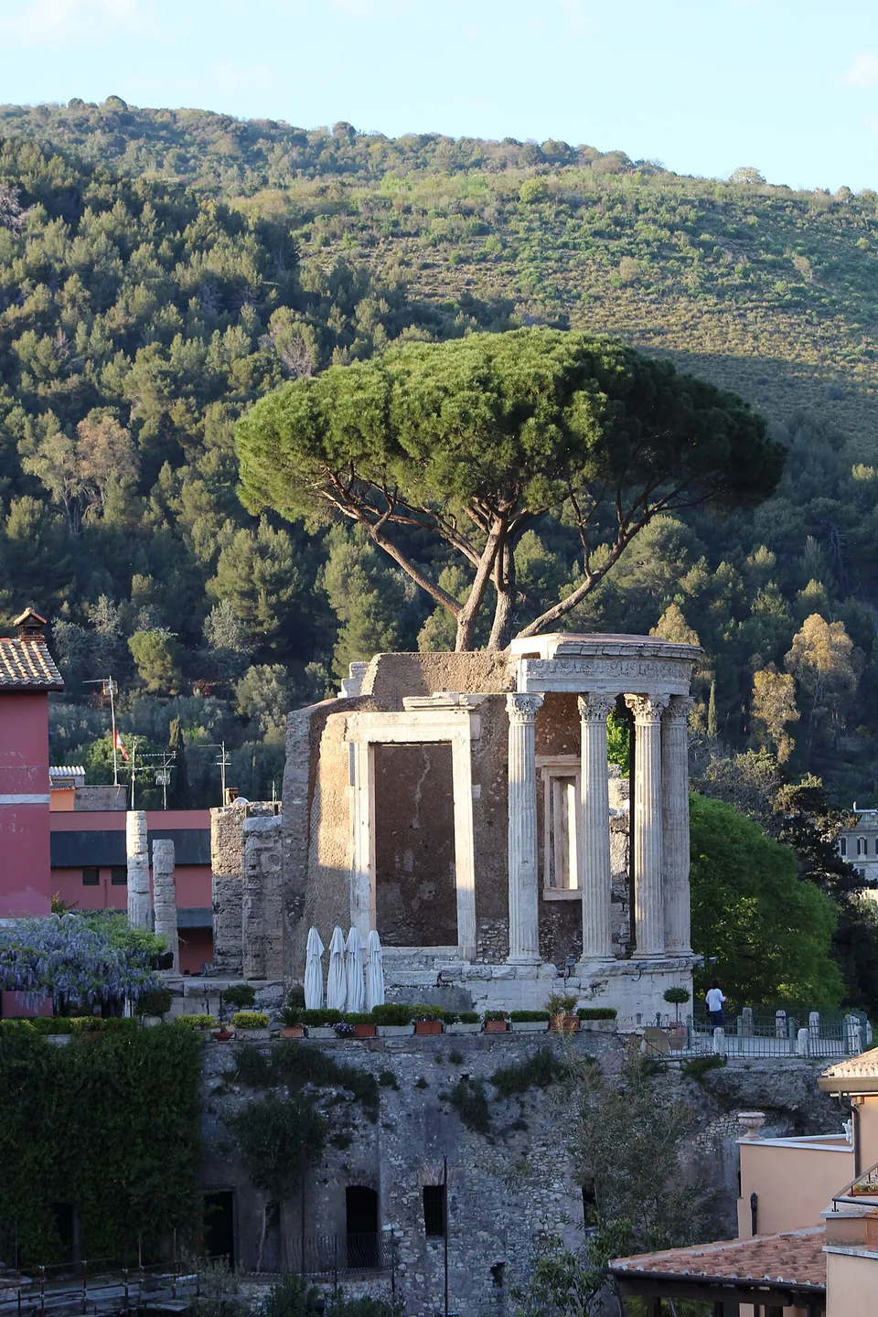 A ruined temple with a tall empty doorway and a row of columns stands on a terrace at the edge of a wooded gorge, houses beside it, a large umbrella pine behind and a forested hillside rising beyond.