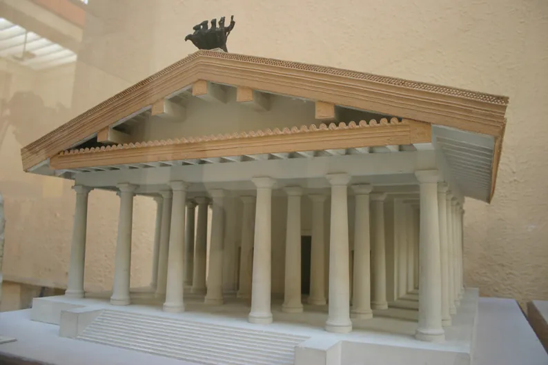 A pale scale model of a broad temple on a high podium with a staircase at the front, rows of columns across the front and down the side, a low pediment and a chariot group on the ridge of the roof.