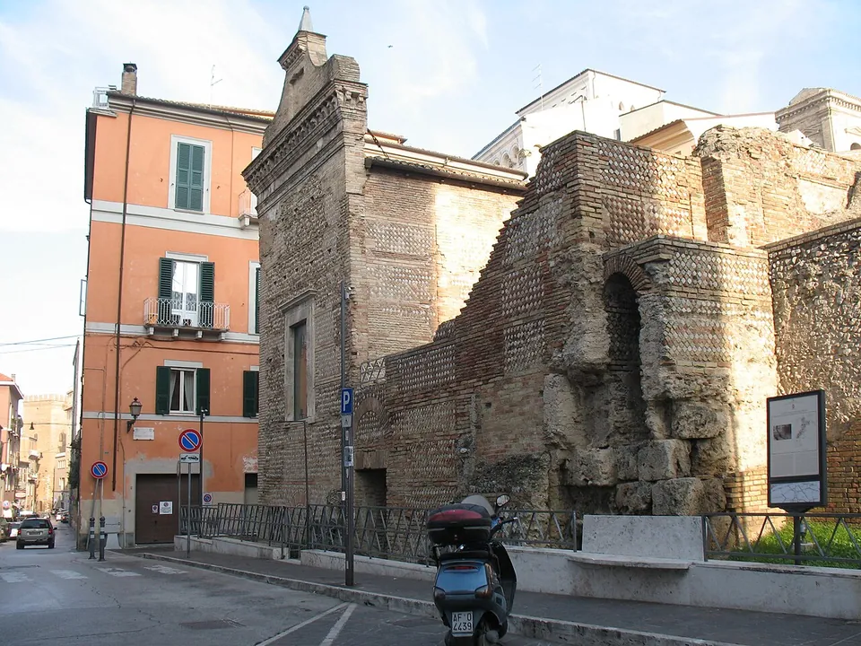 High walls of brick and stone in a town street, with an arched opening low down and a later gabled facade rising at one end, beside a pink apartment building and a parked scooter.