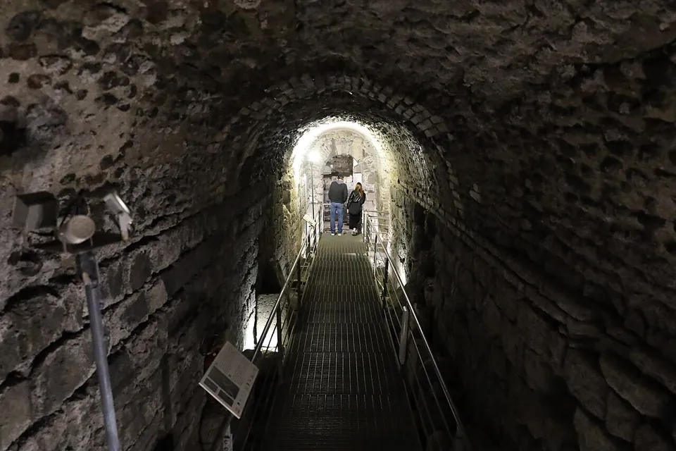 A long, low barrel-vaulted passage of rough stone with a metal grating walkway and handrails running along it, two visitors standing in the light at the far end.