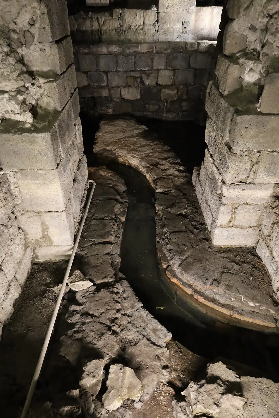 A narrow channel with a smooth curved rim winding across a rough floor between two piers of squared stone, a metal pipe lying along one side.
