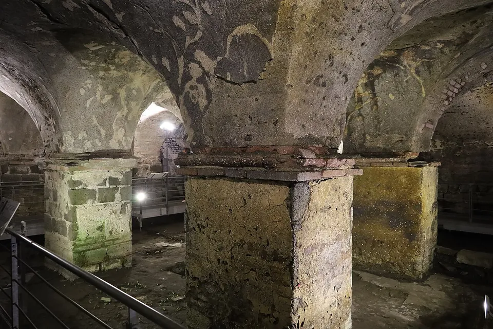 An underground hall lit by floor lamps, its cross-vaulted ceiling springing from thick square pillars of stone blocks, with pale patches of plaster on the dark vaults and a metal railing along a walkway.