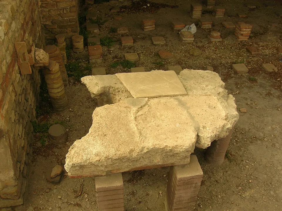 Stacks of square bricks standing in rows on a floor, with a broken slab of mortar and tile resting on some of them and a brick-banded wall at one side.