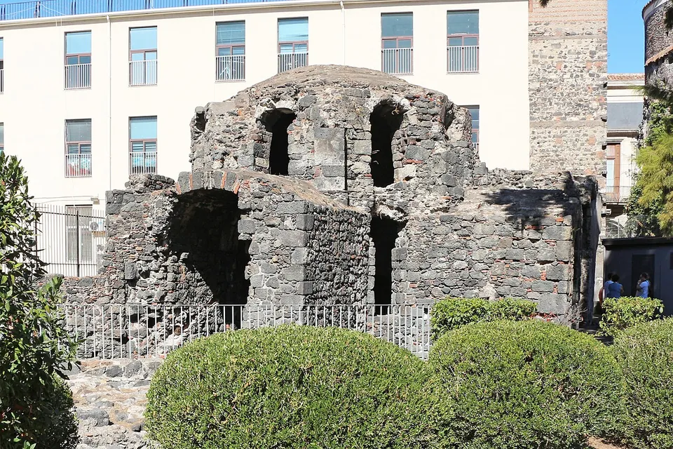 A squat ruined building of dark rubble stone with a low dome and arched openings, standing behind a railing and clipped hedges in front of a modern white block.