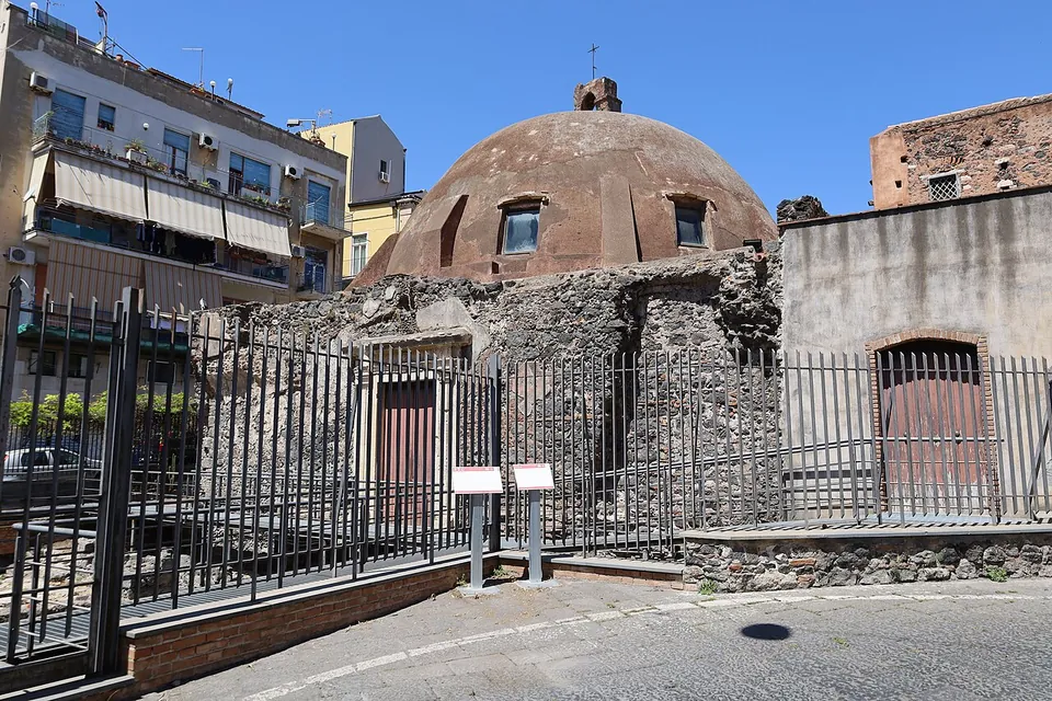 A domed stone building behind a tall iron railing on a city street, with two small information panels standing at the fence and blocks of flats behind.