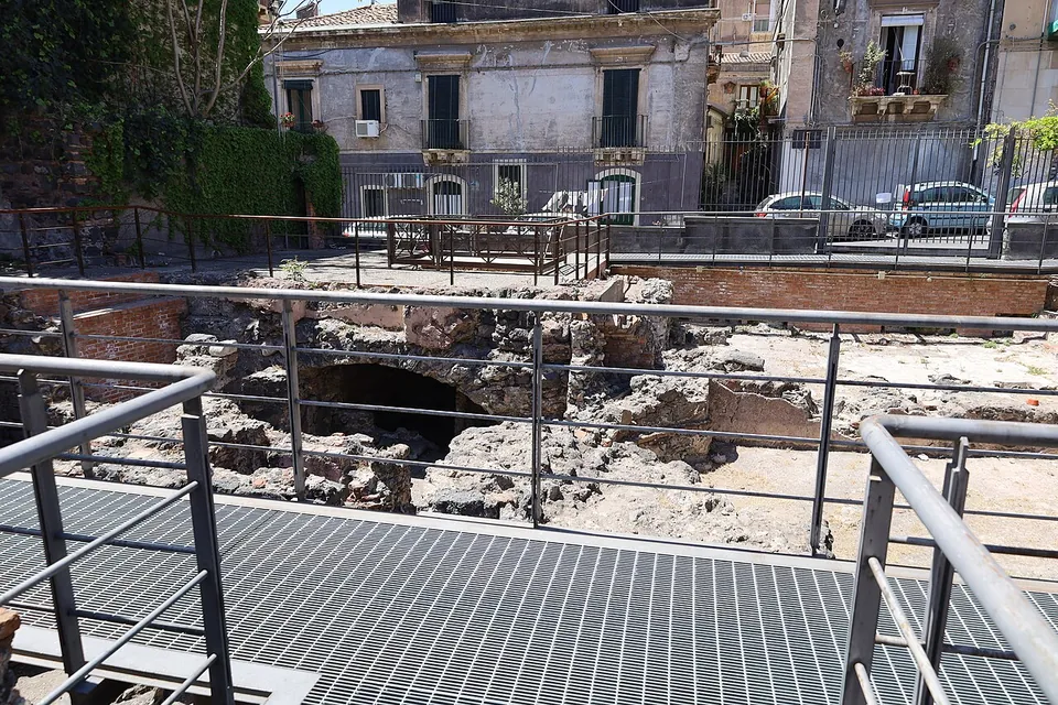 Excavated rubble walls and the dark mouth of a low vaulted chamber seen from a steel grating walkway, with parked cars and buildings beyond the railings.