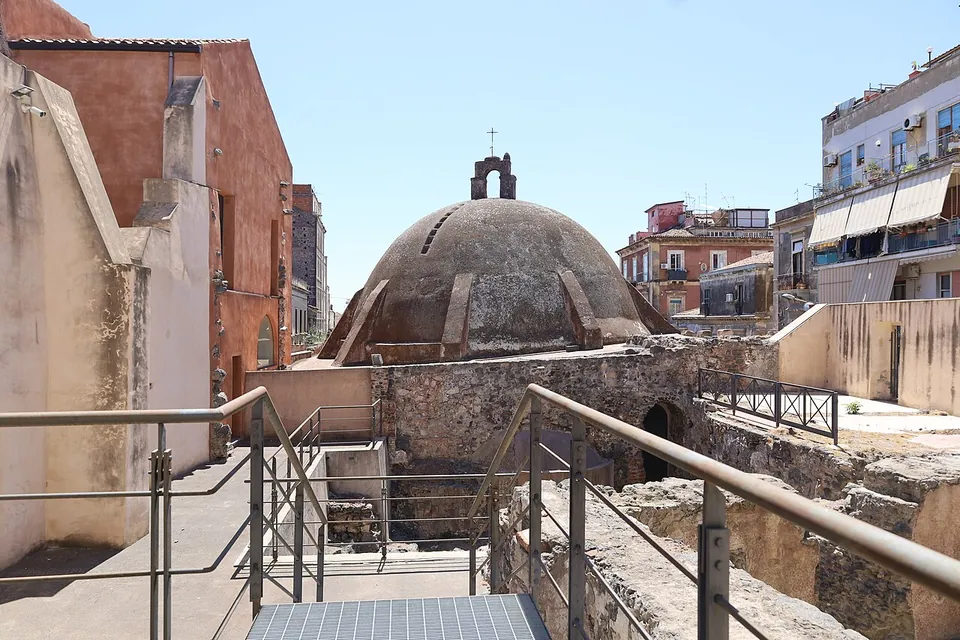 The ribbed dome of the church rising beyond an excavated area of rough stone walls, with metal walkways and handrails leading across it between modern blocks of flats.