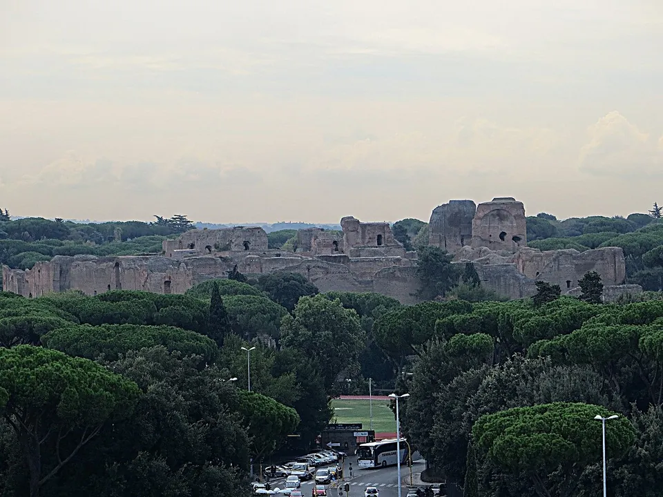A long line of ruined brick walls and tall broken blocks rising above a dense canopy of umbrella pines, seen from a height, with a road, a coach and parked cars in the foreground.