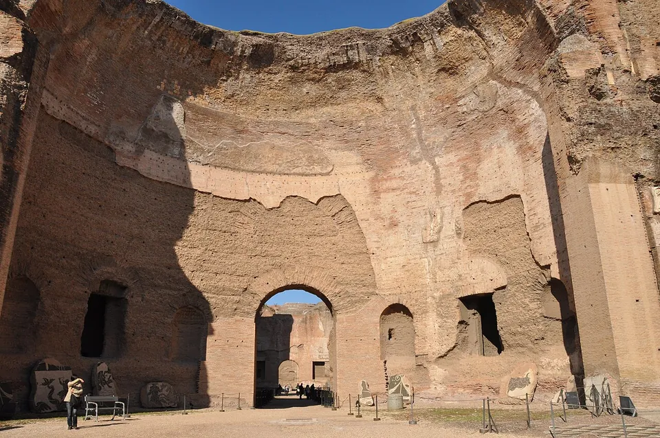 The interior of a vast roofless hall, its brick walls rising high above an arched doorway in the centre, with a few visitors and stone fragments at the foot of the walls.
