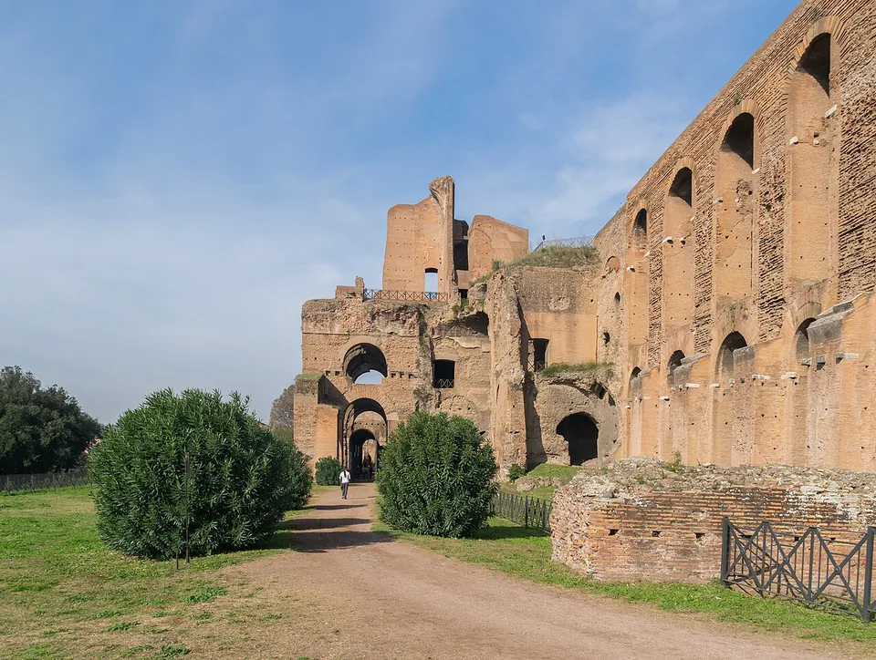 A gravel path leading between rounded shrubs towards high brick walls pierced by rows of arched openings, with a tall ruined brick block rising behind them.
