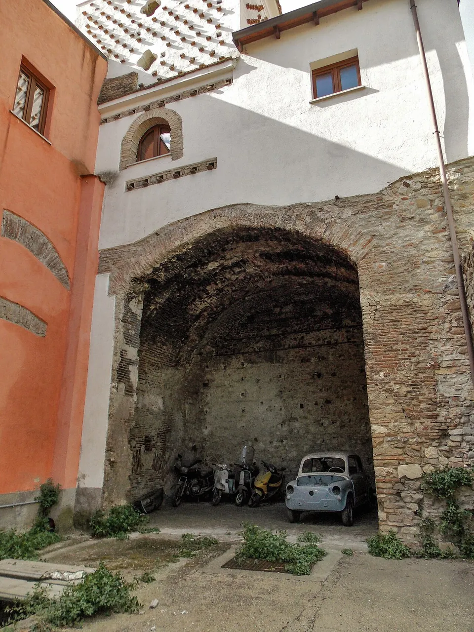A large barrel-vaulted room of brick and stone, open to a courtyard through a broad arch, built into the base of a painted house, with scooters and a small old car parked inside.