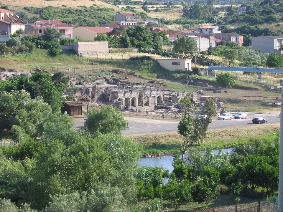 A view from a wooded slope above the river Tirso across to the ruins of the baths, a row of grey stone arches beside a road with a few parked cars, and the houses of Fordongianus on the hillside behind.