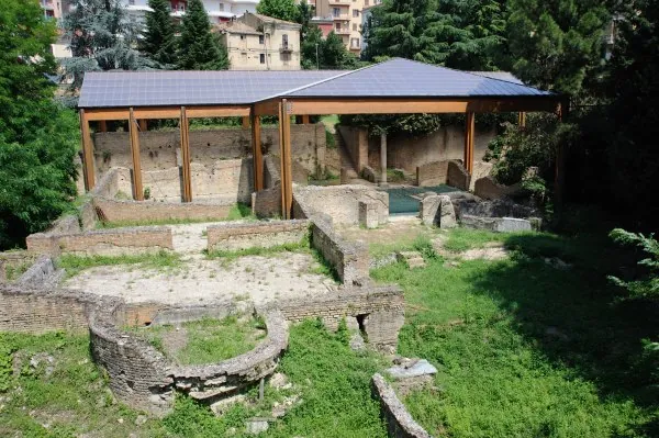 Low brick walls of excavated rooms in the grass, one curving in a semicircle, with a wooden canopy on posts sheltering more walls behind, and apartment blocks above the trees.