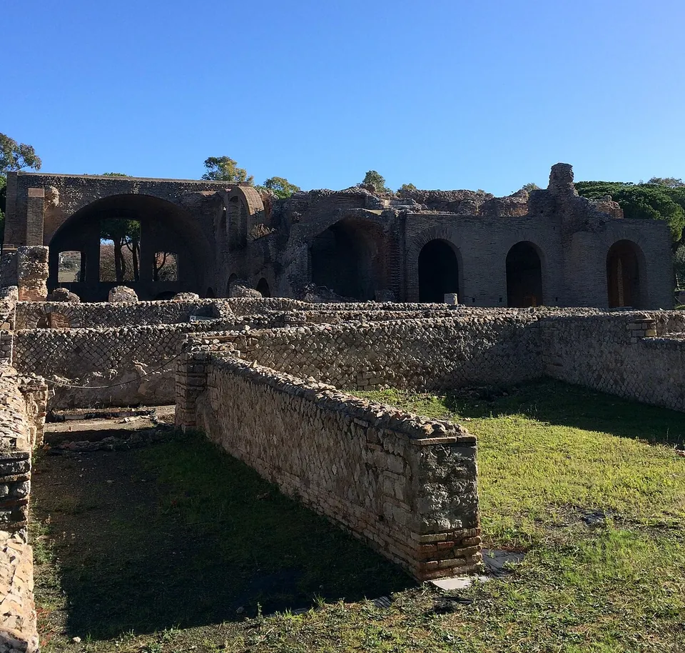 Low brick-faced walls of excavated rooms enclosing a patch of grass in the foreground, and behind them a long brick building with a row of tall round-arched openings, the largest opening onto a vaulted hall.