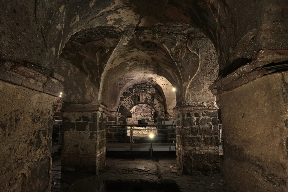 A dimly lit underground hall with cross-vaulted ceilings resting on square stone pillars, with low railings and lights along the floor.