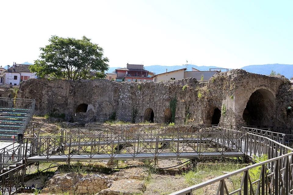 The inside of the amphitheatre, with a raised metal walkway running over excavated ground and, behind it, a curving wall of rough stone pierced by arched openings, modern houses and hills above.