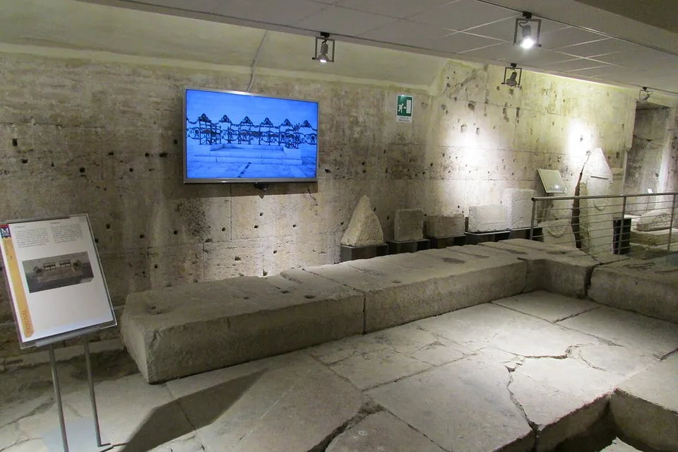 A low platform of large, worn stone blocks on a cracked flagstone floor in a lit underground room, with an information panel on a stand and a screen on the wall showing a drawing of a colonnade.