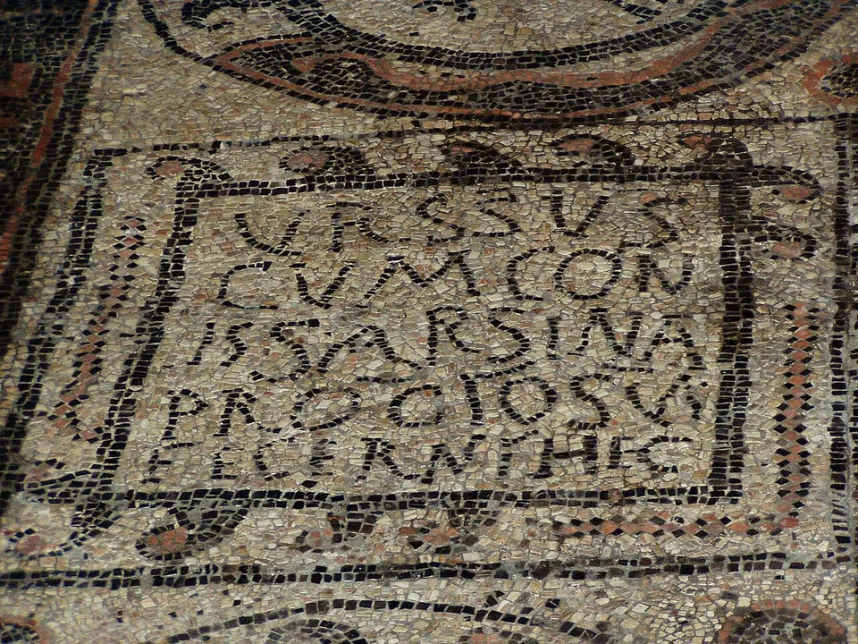 Close view of a mosaic panel framed in dark stones, with five lines of Latin capitals beginning "VRSSVS CVM CON".