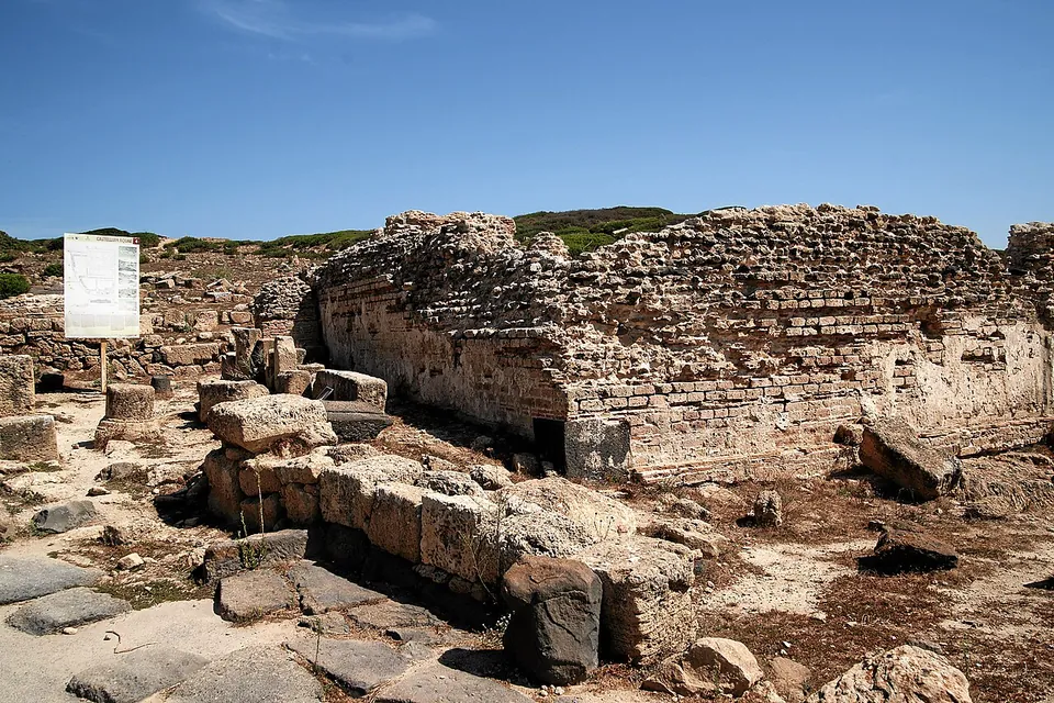 A rectangular block of rubble masonry walls standing above head height behind a row of large cut stones and paving slabs, with an information board on the left.