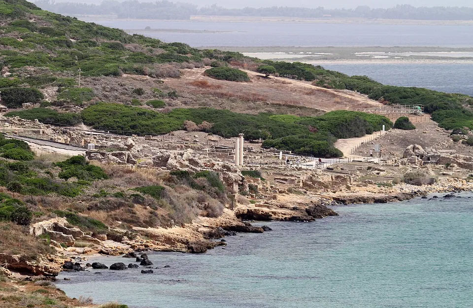 The excavated town spread along a low rocky shoreline below a scrub-covered hillside, with a column standing among the ruins and water and flat land in the distance.