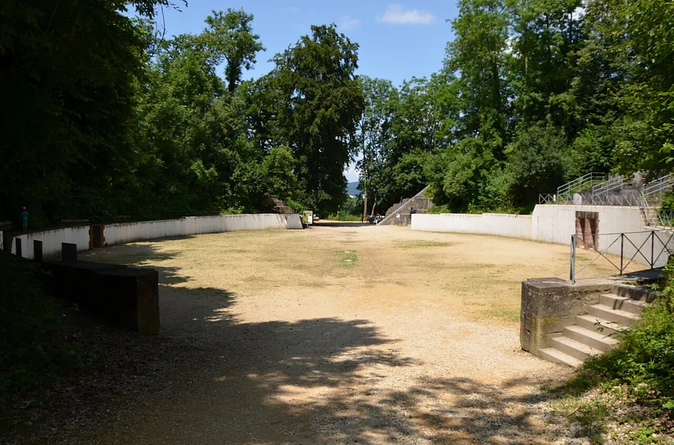 An oval arena of packed earth enclosed by a low white wall, with stone steps leading up the bank on the right and tall trees all around.