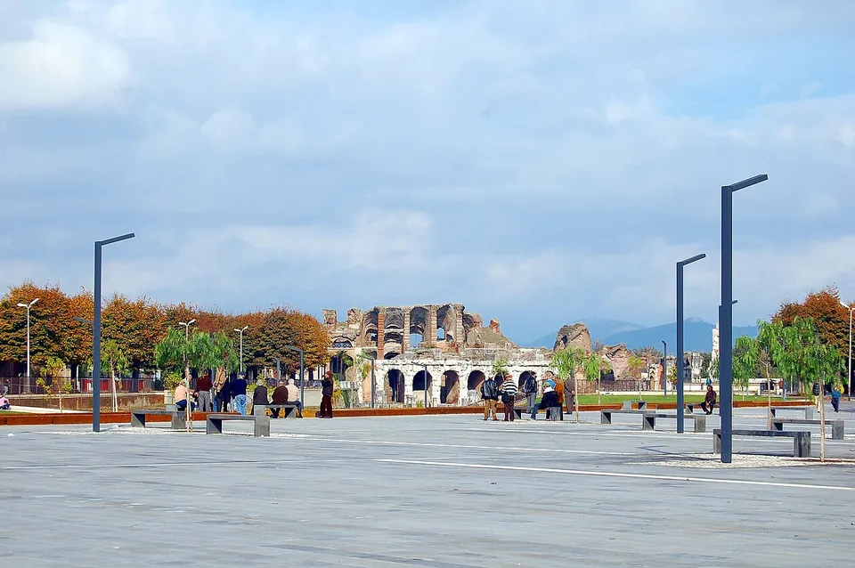 A wide paved square with benches and tall street lamps, people strolling, and beyond a line of autumn trees the ruined arcades of the amphitheatre, with mountains on the horizon.