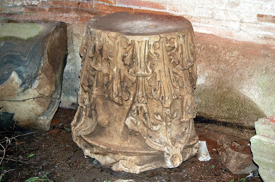 A drum of white marble carved with acanthus leaves, standing on bare earth beside rough stone blocks against a brick wall.