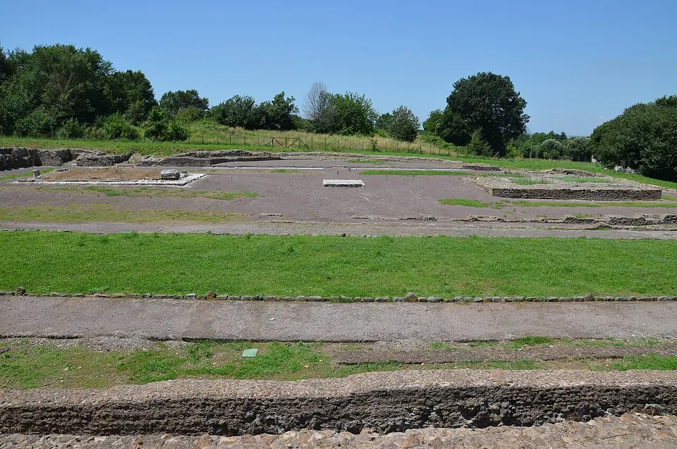 A wide stretch of mown grass crossed by low stone foundations, with the outlines of buildings marked out at ground level and a line of trees behind.