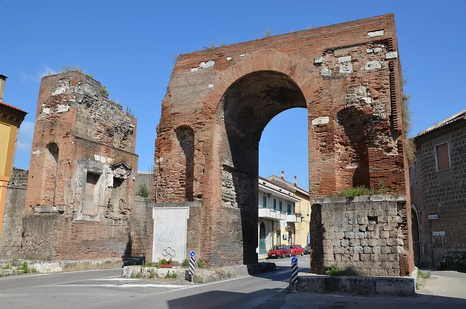 A tall brick arch on a base of stone blocks and, to its left, a free-standing brick pier with niches, standing beside a road among modern houses.