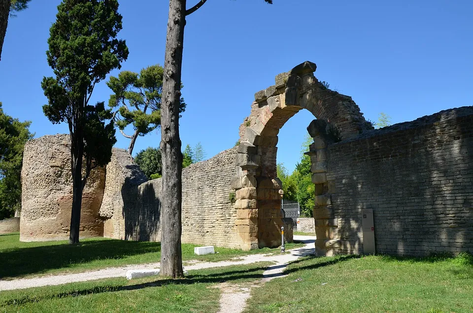 A long wall of small, regular courses of pale stone running across a lawn under pine trees, with the stump of a round tower on the left and a ruined arched gateway of larger blocks breaking the wall line.