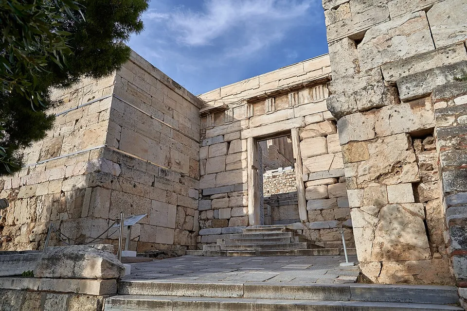 A gateway of large squared stone blocks, a doorway with a stone lintel at the top of a short flight of steps between two plain walls of the same masonry, with an information board on the left.