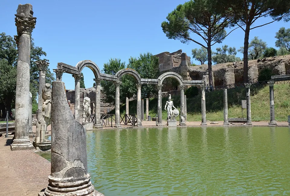 A long rectangular pool of still green water edged with paving, ending in a curved row of columns joined alternately by arches and flat lintels, with statues standing between them and umbrella pines above.