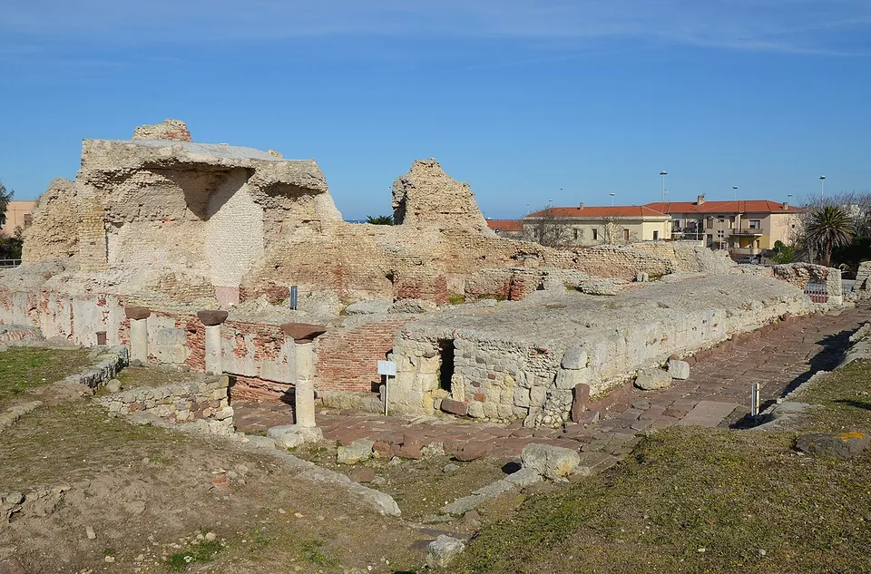 A broad field of excavated ruins under a blue sky, with a tall block of masonry and low walls of stone and brick on the left, a paved street running away to the right and a row of modern houses behind.