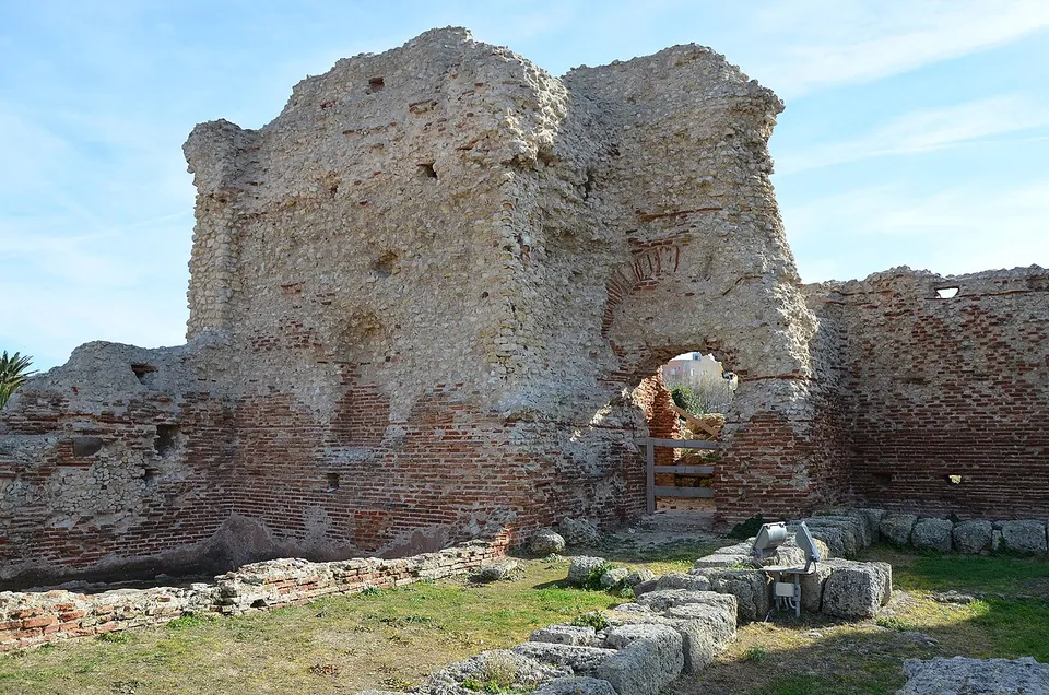 A tall ruined wall of rough rubble core rising above courses of red brick, pierced by an arched doorway, with a low line of stone blocks on the grass in front.