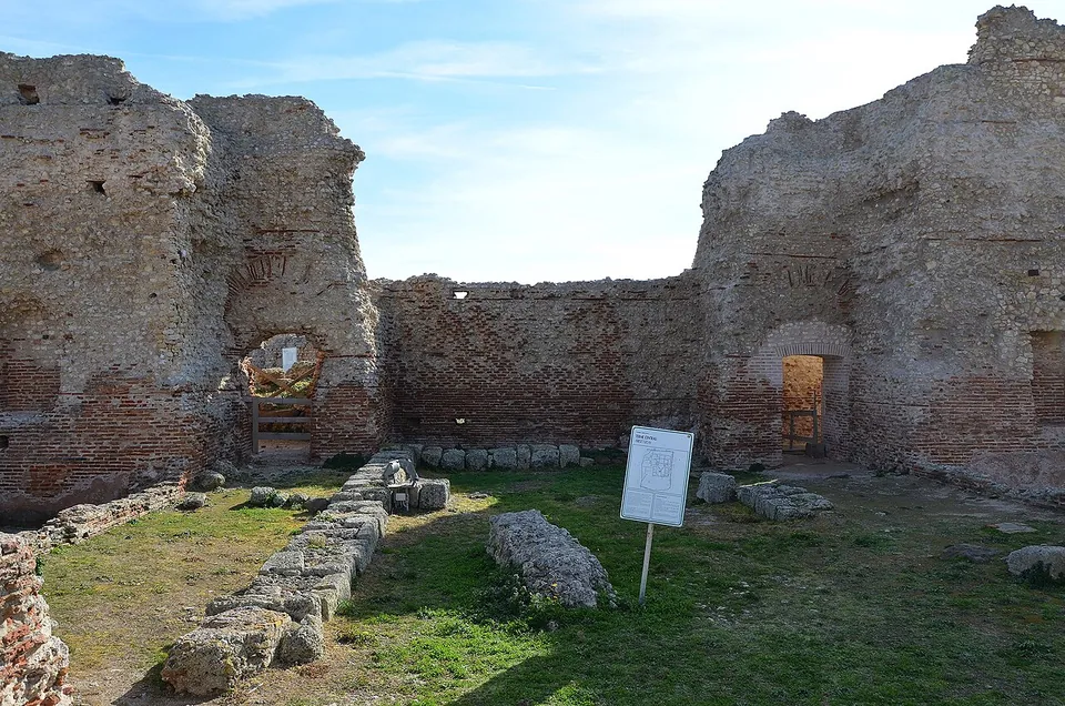 Tall ruined walls of rubble masonry banded with red brick enclose a grassy room, with arched doorways in them, stone blocks lying on the ground and a small information sign in the middle.