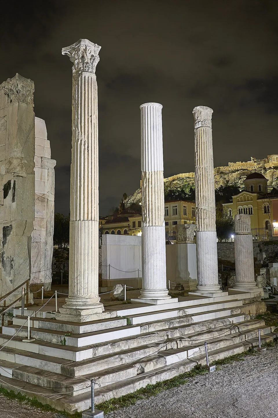 Marble steps leading up to a row of tall fluted columns, two with carved capitals, floodlit at night, with a church and the lit Acropolis rock behind.