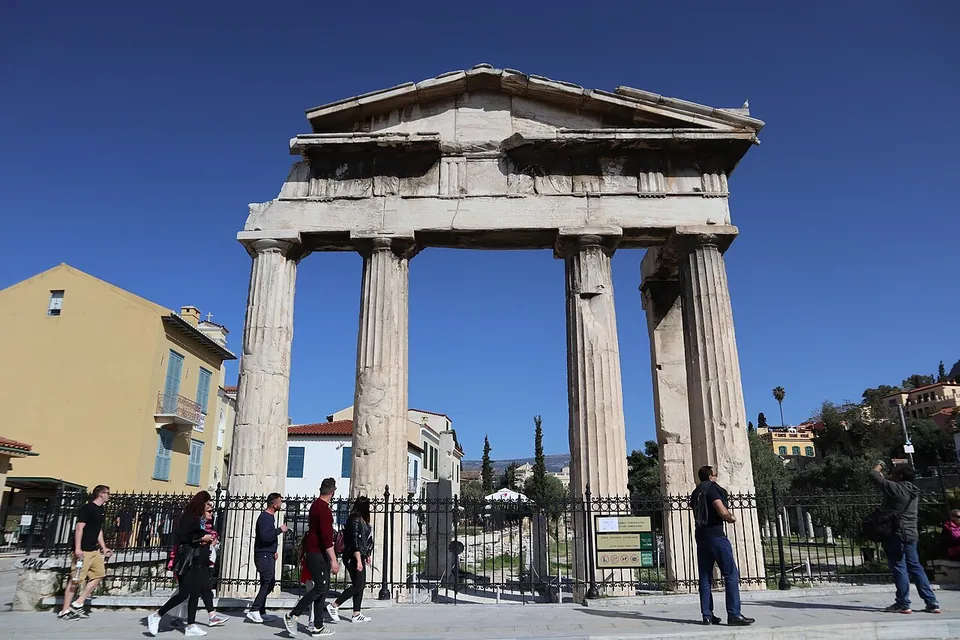 A marble gateway of four thick fluted columns carrying a beam and a low triangular pediment, standing behind a railing on a busy pavement where people walk past.