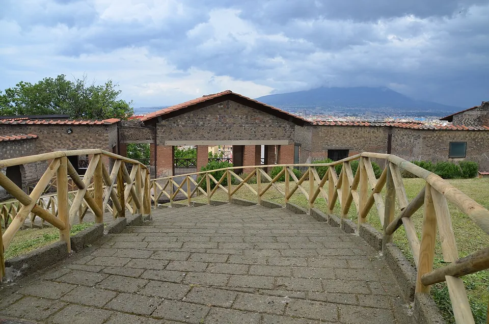 A paved walkway with timber railings curving towards low stone buildings with tiled roofs, with a mountain on the horizon under a cloudy sky.