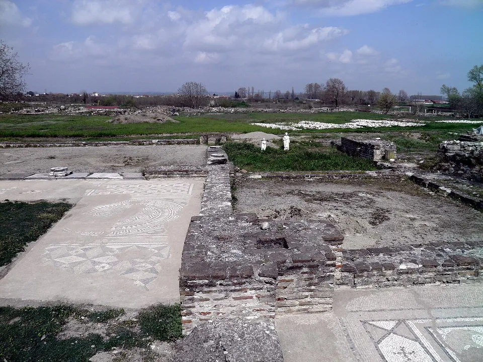 Low brick and stone walls of an excavated building with patterned mosaic floors in the foreground, white statues on a far wall and flat green country stretching away under a cloudy sky.
